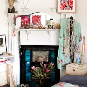May include: A white fireplace mantel with a black fireplace and blue tiles. The mantel is decorated with a variety of items, including a mirror, candles, and a vase of flowers. A light blue robe with floral embroidery hangs on a hook above the fireplace.