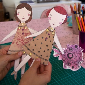 May include: Two paper dolls with brown and black polka dot dresses and floral dresses. The dolls have red and brown hair and are holding a bouquet of purple flowers.
