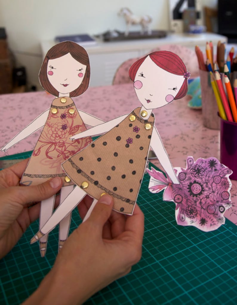 May include: Two paper dolls with brown and black polka dot dresses and floral dresses. The dolls have red and brown hair and are holding a bouquet of purple flowers.
