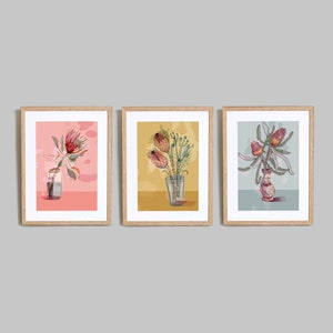 May include: Three framed prints of botanical illustrations featuring pink, red, and yellow flowers in glass vases. The prints are set against a light pink, yellow, and blue background.