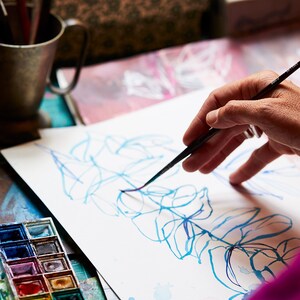 May include: A person's hand uses a paintbrush to draw a blue and purple floral design on a white sheet of paper. A watercolor paint palette with various colors is visible in the foreground.