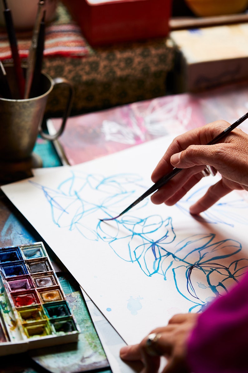 May include: A person's hand uses a paintbrush to draw a blue and purple floral design on a white sheet of paper. A watercolor paint palette with various colors is visible in the foreground.