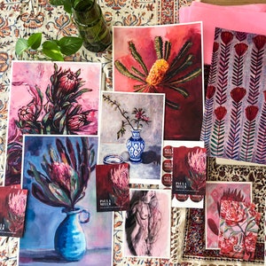 May include: A collection of art prints featuring vibrant floral designs, including proteas and banksias. The prints are displayed on a table with a patterned tablecloth and a pink fabric. The prints are labeled with the artist's name, "Paula Miller".