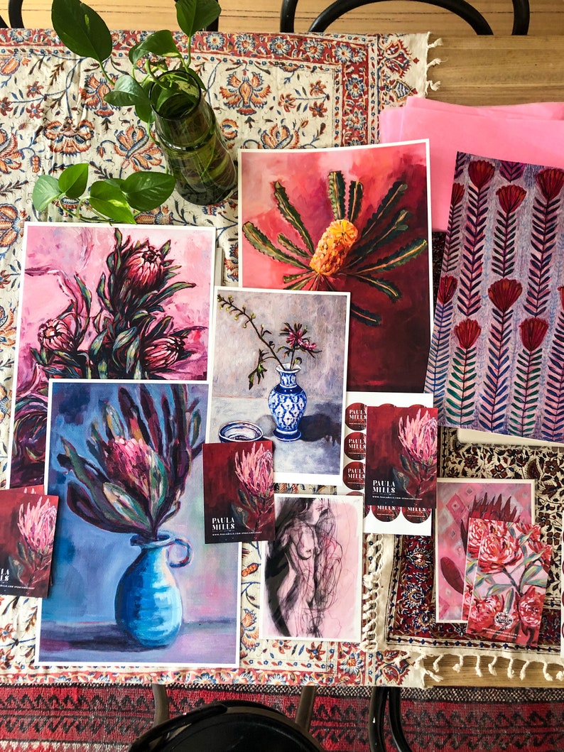 May include: A collection of art prints featuring vibrant floral designs, including proteas and banksias. The prints are displayed on a table with a patterned tablecloth and a pink fabric. The prints are labeled with the artist's name, "Paula Miller".