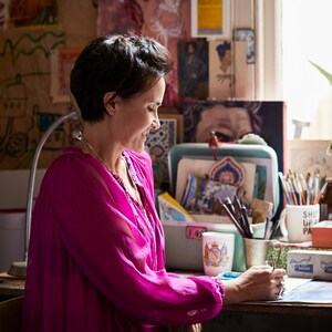 May include: A woman wearing a bright pink blouse sits at a desk with a mug that says "Shut Up and Paint" and a variety of art supplies. She is working on a project with a blue and white design.