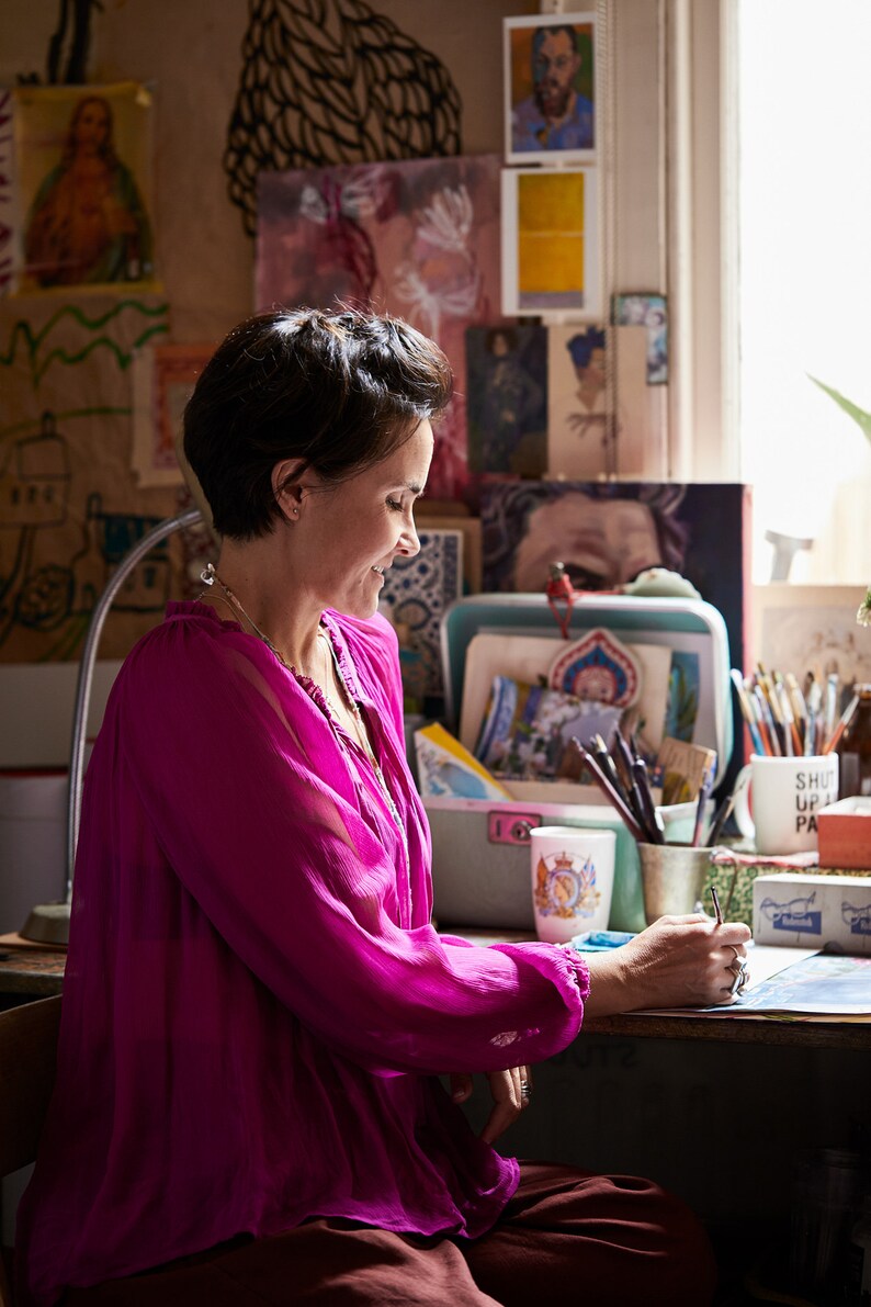 May include: A woman wearing a bright pink blouse sits at a desk with a mug that says "Shut Up and Paint" and a variety of art supplies. She is working on a project with a blue and white design.