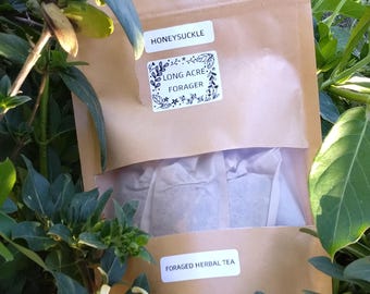 May include: A brown paper bag with a white label that says "Honeysuckle" and "Long Acre Forager" with a floral design. The bag contains several tea bags and says "Foraged Herbal Tea" on the bottom.