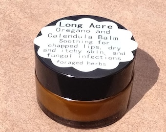 May include: A small glass jar with a black lid containing a brown balm. The label on the jar reads "Long Acre Oregano and Calendula Balm Soothing for chapped lips, dry and itchy skin, and fungal infections foraged herbs."