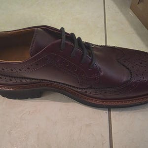 Puede incluir: Zapato tipo wingtip de cuero burdeos con cordones negros. El zapato presenta perforaciones decorativas y una suela marrón. Diseñado para vestimenta formal o de negocios.