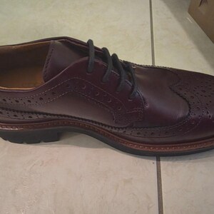Puede incluir: Zapato Oxford de cuero burdeos con cordones negros. El zapato presenta perforaciones decorativas y una suela marrón. Diseñado para vestimenta formal o de negocios.