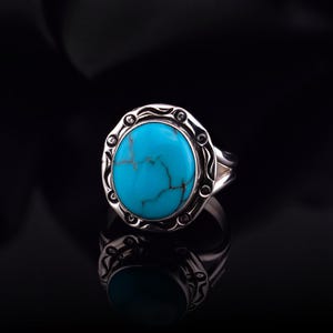 May include: A silver ring with a large, oval turquoise stone. The stone has brown veining and is set in a decorative silver bezel. The ring is displayed on a reflective black surface.