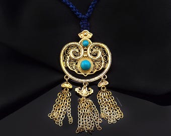 May include: Gold-toned pendant necklace with a filigree design, featuring three turquoise-colored stones. The pendant has three dangling chain tassels and is suspended from a blue cord.