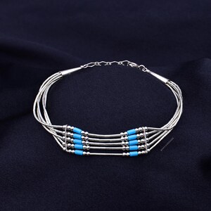 May include: Silver-toned bracelet with multiple strands, featuring turquoise-colored beads. The bracelet has a delicate chain closure and is displayed on a dark blue fabric, showcasing a minimalist design.