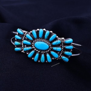 Puede incluir: Un brazalete de plata con una piedra turquesa ovalada central y piedras turquesas más pequeñas. Las piedras están engastadas en un engaste de plata detallado con un diseño de cuerda trenzada. El brazalete tiene un diseño curvo.
