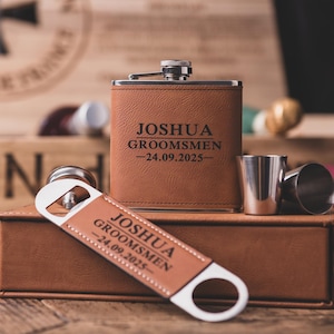 May include: A brown leather hip flask, bottle opener, and shot glasses set. The flask and bottle opener are engraved with "JOSHUA GROOMSMEN -24.09.2025-". The set is displayed on a wooden surface.
