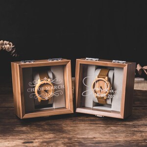 May include: Two wooden watches in display boxes. The watches have light brown leather straps and wooden faces. The boxes are made of wood and have glass fronts. The text "Chloe March" and "Jordan 18.06.2020" are engraved on the glass.