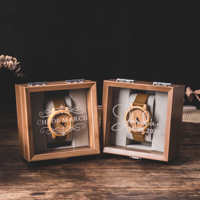 May include: Two wooden watches in display boxes. The watches have light brown leather straps and wooden faces. The boxes are made of wood and have glass fronts. The text "Chloe March" and "Jordan 18.06.2020" are engraved on the glass.
