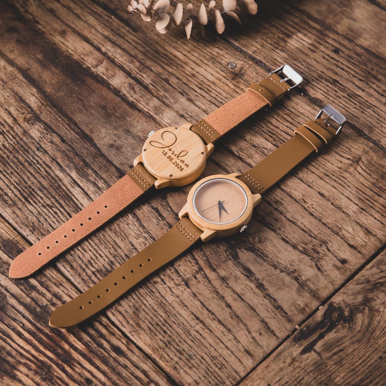 May include: Two wooden watches with brown leather straps are displayed on a wooden surface. One watch has the name "Jordan" and the date "18.08.2026" engraved on the face. The other watch has a simple wooden face with black hands.