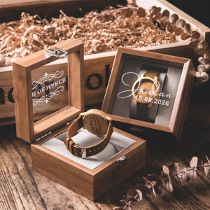 May include: Wooden watch with a brown leather band displayed in a bamboo box. The watch face is visible, and the box has a clear glass top. The watch is engraved with text. Another box displays a watch with the name "Jordan" and the date "18.08.2026".