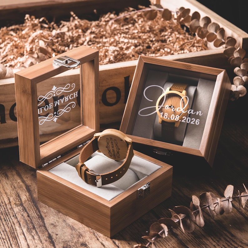 May include: Wooden watch with a brown leather band displayed in a bamboo box. The watch face is visible, and the box has a clear glass top. The watch is engraved with text. Another box displays a watch with the name "Jordan" and the date "18.08.2026".