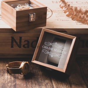 May include: Wooden watch box with a glass top and a silver clasp. The box is engraved with the name "CHLOE MARCH". A wooden watch with a leather band is next to the box. The watch box is 6 inches long.