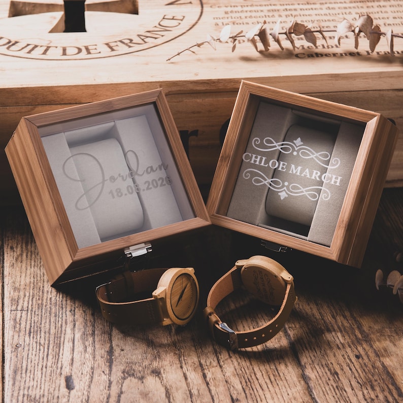 May include: Two wooden watch display boxes with glass tops, each personalized with a name and date. Two wooden watches with brown leather straps are displayed next to the boxes. The boxes and watches are on a wooden surface.