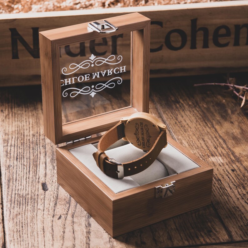 May include: A wooden watch with a brown leather strap rests inside an open wooden box with a glass lid. The watch face is visible, and the box is lined with a light-colored fabric. The box has a silver clasp.