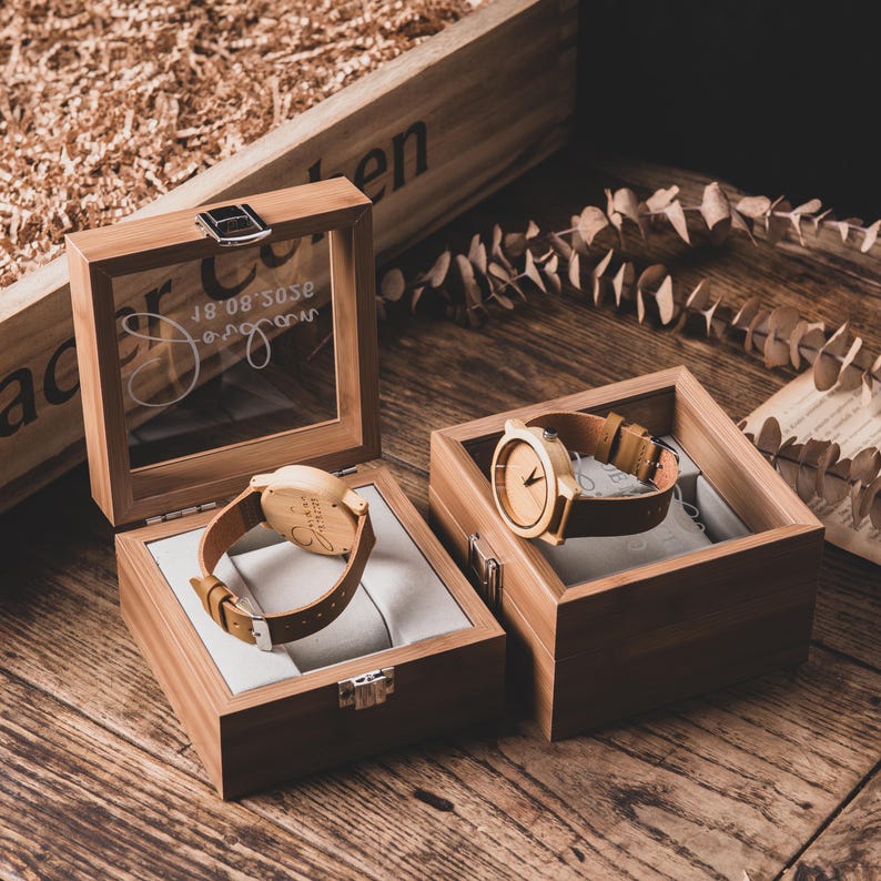 May include: Wooden watches with leather straps displayed in wooden boxes. The watch faces and boxes are light brown. The watch straps are brown. One box is open, revealing the watch inside. The other box is closed with a glass top.