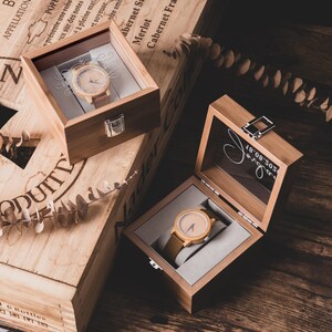 May include: Two wooden watches with light brown faces and brown leather straps are displayed in open wooden boxes. The boxes are on a wooden surface, with text on the boxes. One box has a silver clasp.
