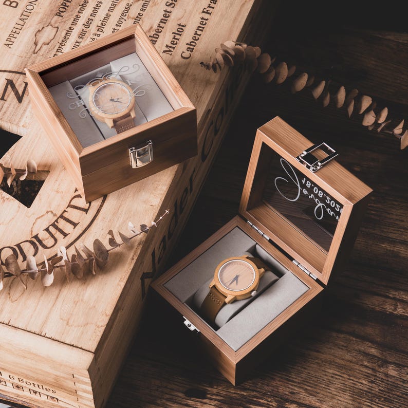 May include: Two wooden watches with light brown faces and brown leather straps are displayed in open wooden boxes. The boxes are on a wooden surface, with text on the boxes. One box has a silver clasp.