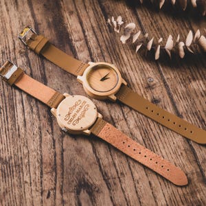 May include: Two wooden watches with tan leather straps are displayed on a wooden surface. One watch has a visible watch face, while the other has the text "CHLOE MARCH" engraved on the back. The watches have a natural wood color.