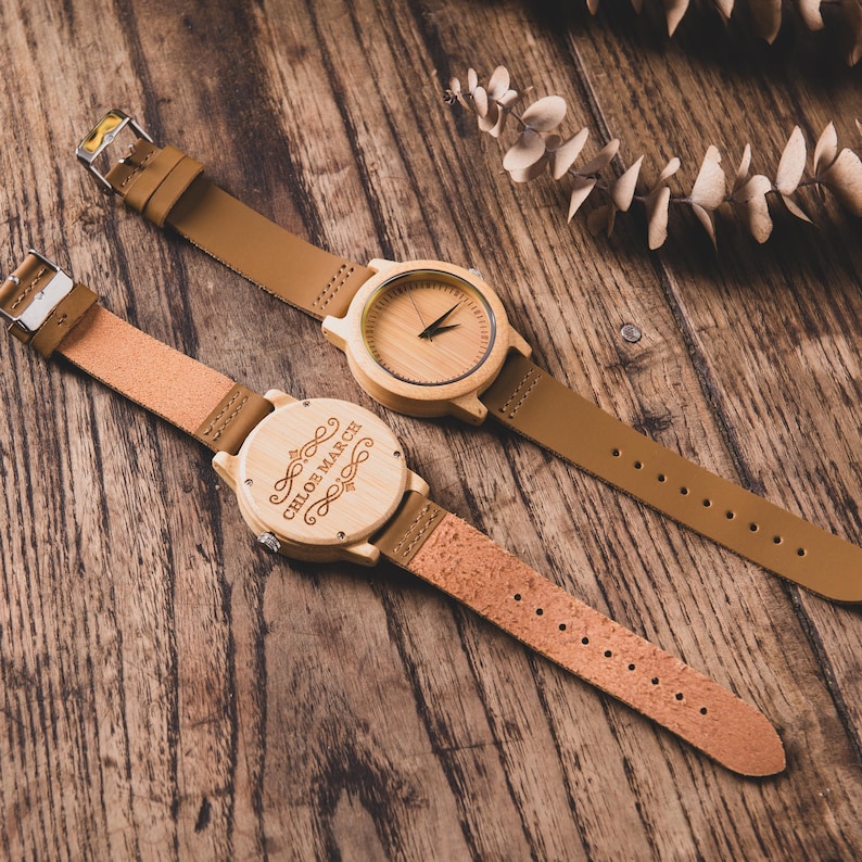 May include: Two wooden watches with tan leather straps are displayed on a wooden surface. One watch has a visible watch face, while the other has the text "CHLOE MARCH" engraved on the back. The watches have a natural wood color.
