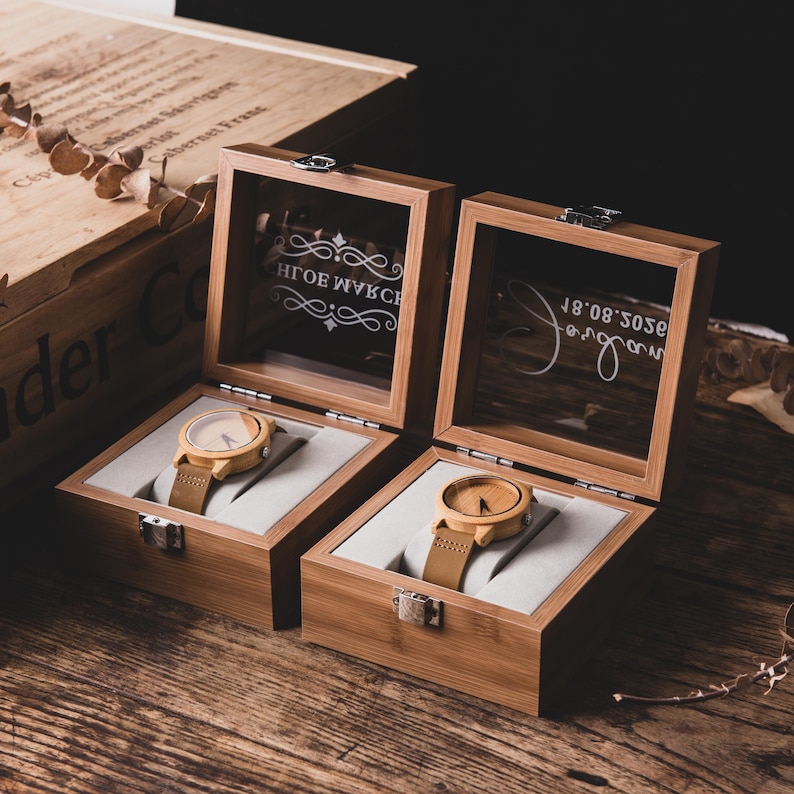 May include: Two wooden watches with light brown leather straps are displayed in open wooden boxes with glass lids. The watch faces are light-colored, and the boxes sit on a wooden surface. The boxes have a light-colored interior.