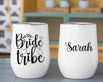 May include: Two white stainless steel stemless tumblers with black lettering. The left tumbler features the text Bride tribe with a diamond ring and heart icon. The right tumbler displays the name Sarah in a cursive black script font.