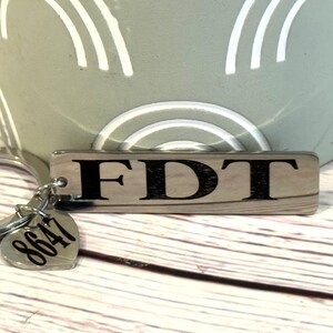 May include: A wooden keychain with the letters "FDT" in black. The keychain has a silver ring and a heart-shaped tag with the number "8647". The background includes a light green object with white arched lines.