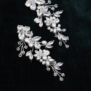May include: A pair of silver bridal hair clips with crystal and pearl embellishments. The clips are designed in a cascading floral pattern.