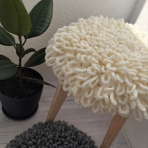 Könnte beinhalten: Ein weißer, flauschiger, runder Hocker aus Strick mit Holzbeinen. Ein grauer, flauschiger, runder Hocker aus Strick befindet sich im Vordergrund.