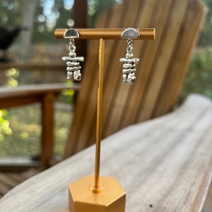 May include: Silver-toned Inukshuk earrings displayed on a gold-colored stand. Each earring features a small, stacked stone design hanging from a half-circle stud. The earrings are presented against a blurred outdoor background.