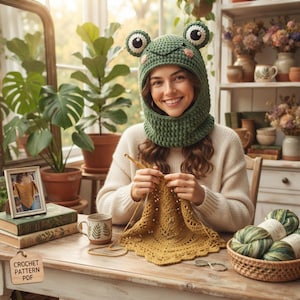 May include: A green crochet balaclava featuring a frog design with large white and black eyes and pink cheeks. The item is worn while working on a mustard yellow crocheted piece with a wooden hook. A wooden tag reads Crochet Pattern PDF.