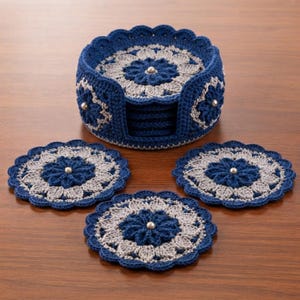 Puede incluir: Un juego de posavasos redondos de ganchillo en hilo azul marino y gris claro, con un patrón floral y una cuenta metálica central. El juego incluye un soporte cilíndrico de ganchillo a juego con un borde festoneado para guardarlos.