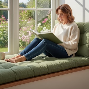 Puede incluir: Una persona lee un libro sentada en un cojín verde junto a una ventana. Viste un suéter color crema y vaqueros azules. Una taza está cerca. La ventana da a un jardín con flores rosas.
