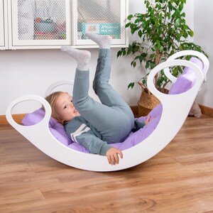 Op de afbeelding: Een wit houten wiebelbord met een paars kussen. Een kind ligt op het kussen met de benen in de lucht.