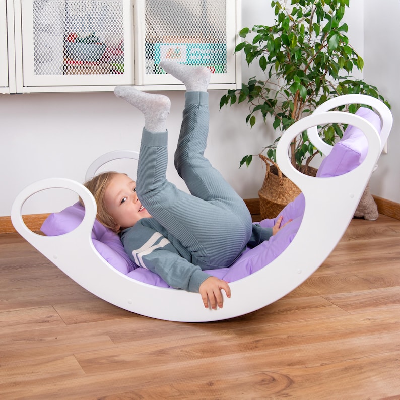 Op de afbeelding: Een wit houten wiebelbord met een paars kussen. Een kind ligt op het kussen met de benen in de lucht.