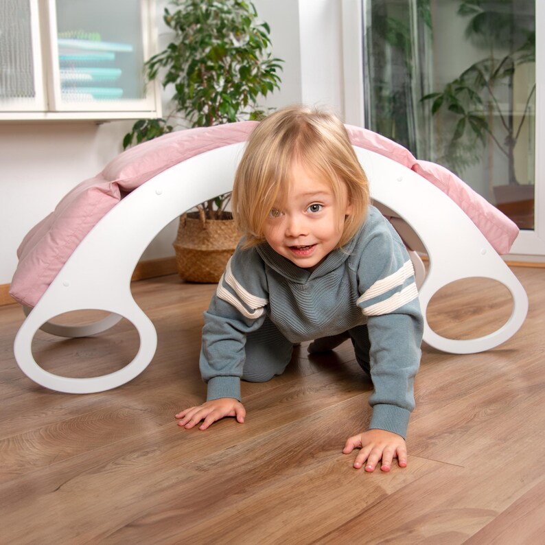 Op de afbeelding: Een witte houten boog met een roze kussen erop. Een klein kind kruipt door de boog.