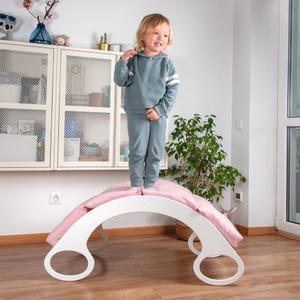 Op de afbeelding: Een witte houten boog met afgeronde randen en twee grote gaten aan beide zijden. De boog is bedekt met een roze deken en een kind staat erop.