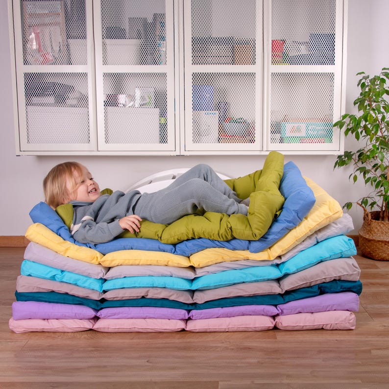 May include: A stack of colorful, rectangular cushions, some with a quilted texture, arranged in a pyramid shape. A young child is lying on top of the cushions, smiling.