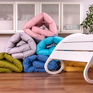 May include: A stack of colorful, folded cushions in various shades of pink, gray, blue, green, and yellow. The cushions are arranged in a pyramid shape, with a white wooden rocking chair in the foreground.
