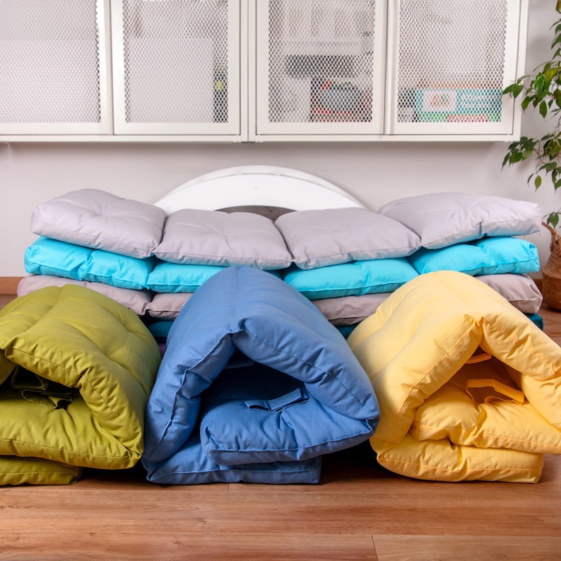 May include: Four folded futon mattresses in different colors: green, blue, yellow, and gray. The mattresses are arranged in a row on a wooden floor.