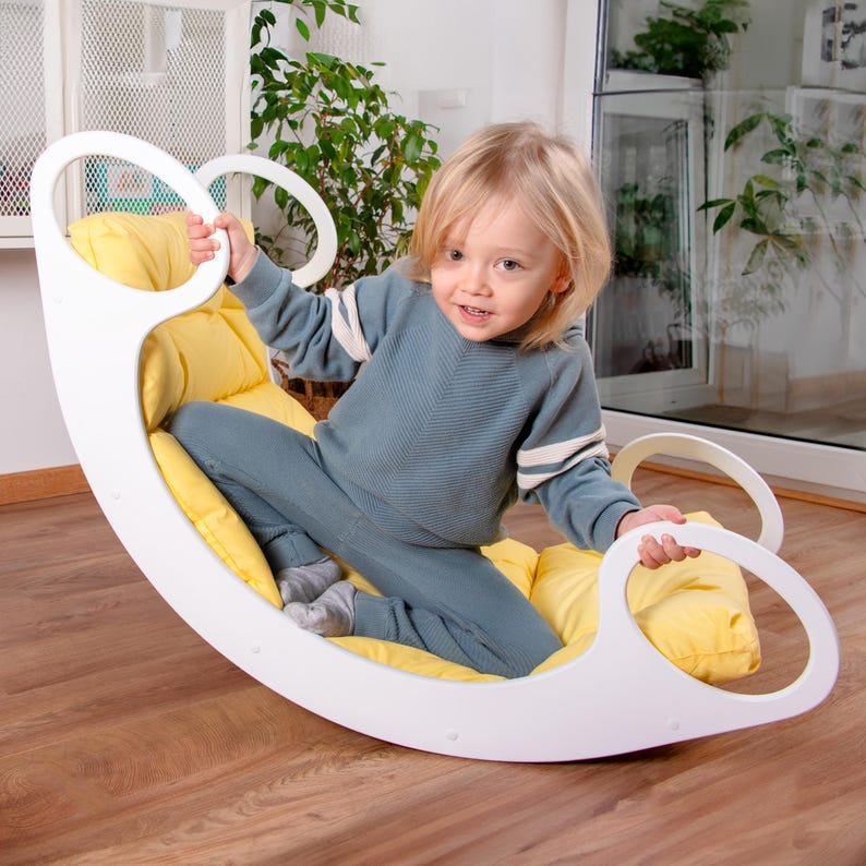 Op de afbeelding: Een witte houten schommelstoel met een geel kussen. Een kind zit in de stoel en houdt de handvatten vast.