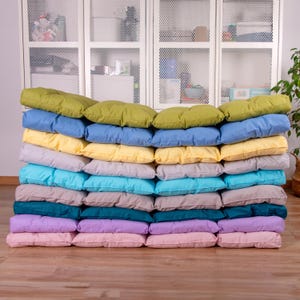 May include: A stack of 15 colorful cushions in various shades of green, blue, yellow, turquoise, gray, teal, purple, and pink. The cushions are arranged in a neat pile, with the top cushion being green.